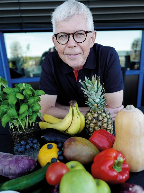 Mann med briller lener seg p? bord. P? bordet ligger frukter som banan, papirka og agurk.  
